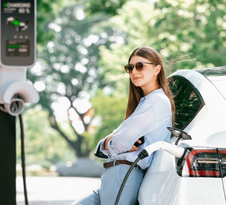 Young woman recharging battery for electric car during road trip travel EV car in natural forest or national park. Eco friendly travel during vacation and holiday. Exalt